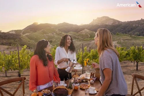 Three women enjoy outdoor dining at a vineyard, featuring fresh food, drinks, and a scenic mountain backdrop at sunset.
