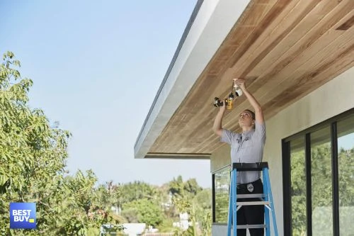 A woman stands on a ladder outdoors and installs recessed lighting in a porch ceiling, with a drill in her right hand, under blue sky.