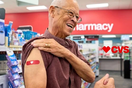 An elderly man laughs in a drugstore, wearing a band-aid with a heart on his arm after vaccination. Pharmacy and logo visible.
