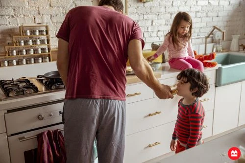 A man prepares food in a kitchen, offering a bite to a young boy while a girl sits nearby. Natural light and a homey atmosphere.