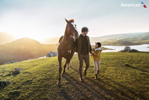 Adult man and child walking with a horse on a grassy hilltop with a mountain vista, lake, and bright sun in the background.