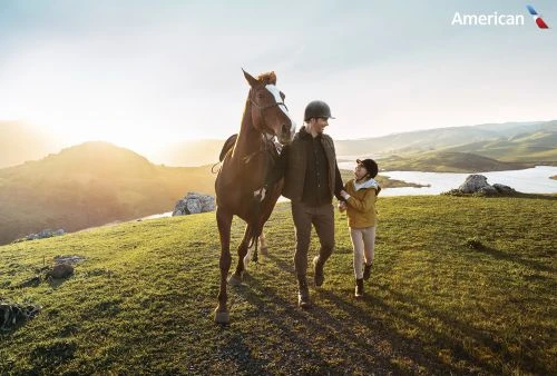 Adult man and child walking with a horse on a grassy hilltop with a mountain vista, lake, and bright sun in the background.