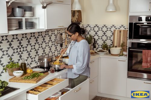 Woman cooking in a modern kitchen with white cabinets, stainless steel appliances, and geometric patterned backsplash tiles.