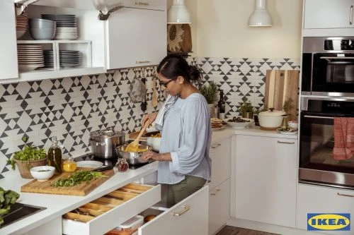 Woman cooking in a modern kitchen with white cabinets, stainless steel appliances, and geometric patterned backsplash tiles.