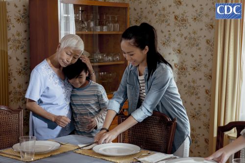 Three generations of an Asian family at a set table: a young woman setting the table, a boy, and an elderly woman with short white hair.