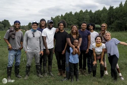 A diverse group of twelve people standing together in a grassy field with trees in the background, photographed by Augustus Butera.