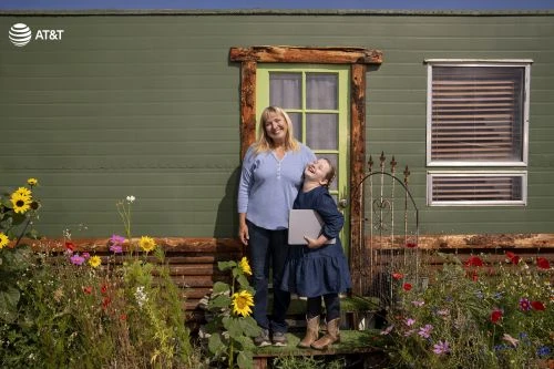 Smiling adult woman and young girl stand outside a green home surrounded by flowers, holding a laptop, captured in a medium shot.