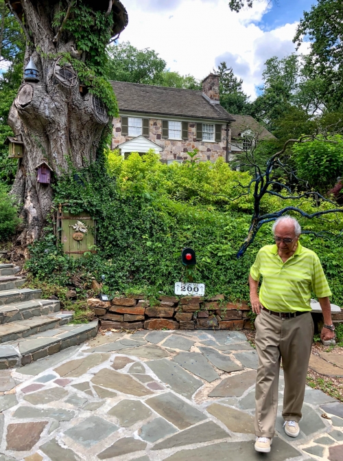 Elderly man walks on a stone path near a large tree with birdhouses, and a stone house is visible in the background on a sunny day.