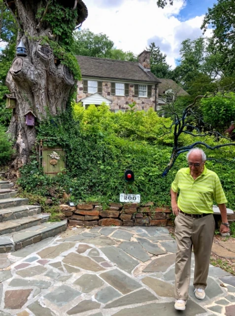 Elderly man walks on a stone path near a large tree with birdhouses, and a stone house is visible in the background on a sunny day.