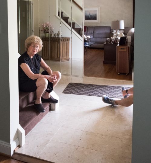 Elderly woman in a black dress sits on the stairs in a brightly lit foyer with light marble floors and mirrored wall in the background.