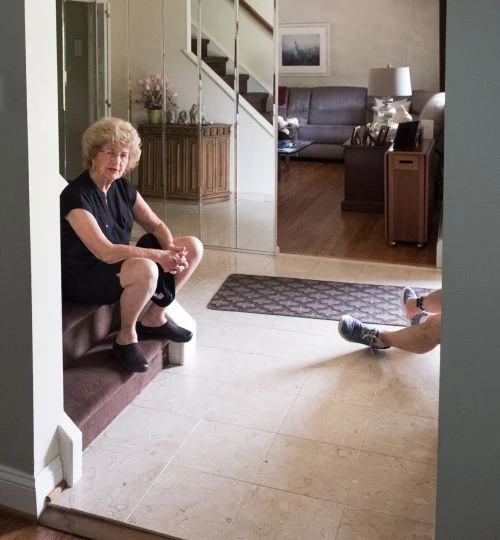 Elderly woman in a black dress sits on the stairs in a brightly lit foyer with light marble floors and mirrored wall in the background.
