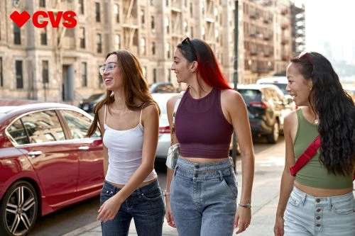 Three young women walk on an urban sidewalk past parked cars and apartment buildings in natural light, wearing casual summer clothing.