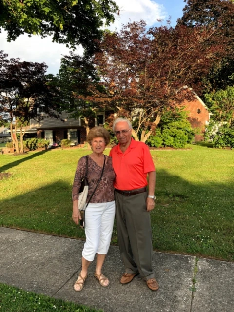 An elderly couple poses on a sidewalk in front of a green lawn and a brick house on a sunny day, creating a warm and inviting scene.