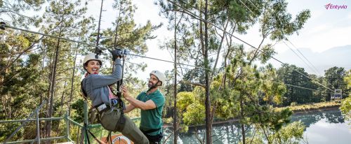 A smiling woman in a harness and helmet rides a zip line, assisted by a man on a platform, with lush trees and mountains in the background.