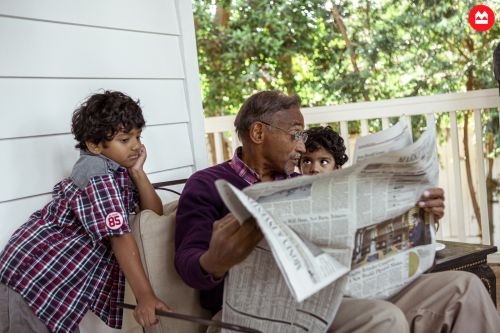 Man and two children read newspaper on porch. Older man in eyeglasses sits while two young children look on at printed news.