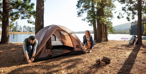Man and woman camping near a lake setting up a tent on a sunny day surrounded by trees and forest in a nature and wildlife setting.