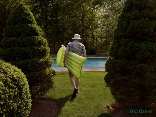 Man in hat walks on lawn toward pool, carrying striped inflatable raft between evergreen trees, Citizens logo in corner.