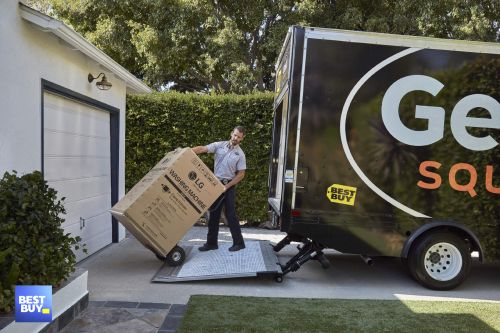 Adult male delivery worker unloads a boxed LG washing machine from a truck ramp outside a house; Best Buy logo is visible.