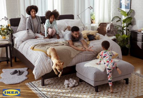 Family of four plays on a modern bed in a naturally lit bedroom with white walls, wood floors, decorative rug, and IKEA branding.