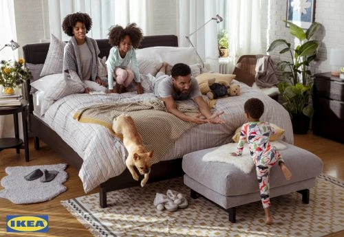 Family of four plays on a modern bed in a naturally lit bedroom with white walls, wood floors, decorative rug, and IKEA branding.