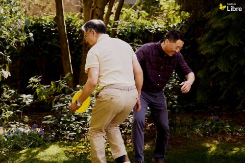 Two Asian men playing frisbee in a lush green garden on a sunny day. One man throws the yellow disc while the other watches.