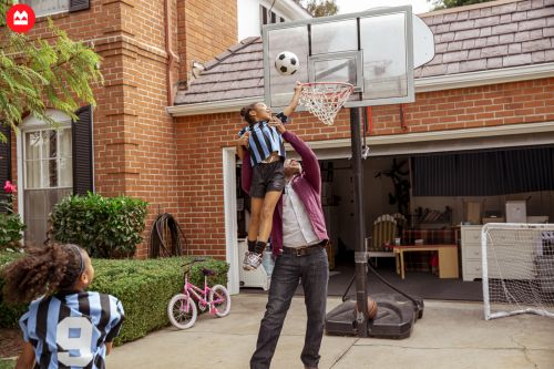 Father and daughter playing basketball in the driveway. The daughter is on his shoulders, reaching for the ball near the hoop.
