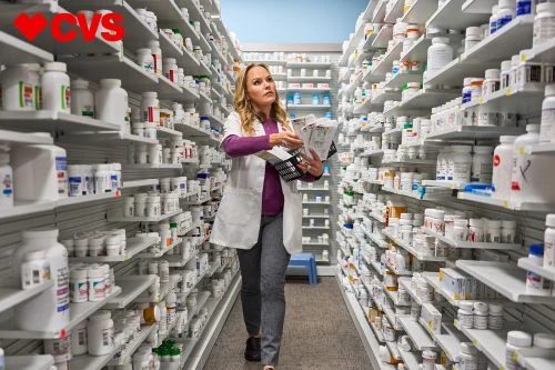 Female pharmacist walking through the aisle of a drugstore, holding a basket with medicine boxes on shelves stocked with products.