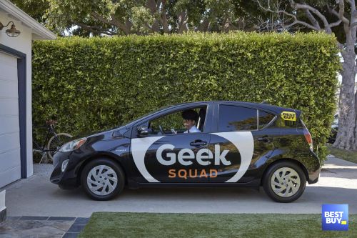 Geek Squad car parked in a driveway with a technician visible through the window; lush green foliage background; bicycle in garage.