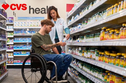Man in a wheelchair reviews product with pharmacy worker at CVS aisle with various vitamins and supplements in natural indoor light.