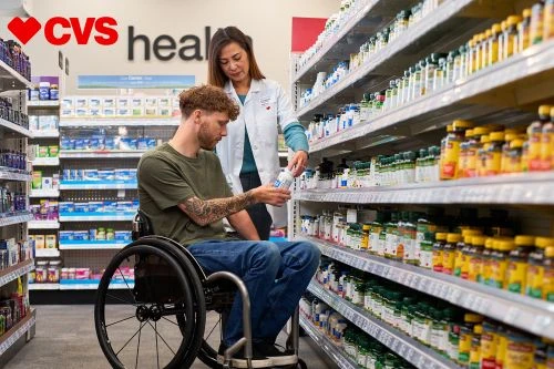 Man in a wheelchair reviews product with pharmacy worker at CVS aisle with various vitamins and supplements in natural indoor light.