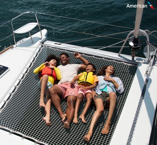 Family of four relaxes on a catamaran over the sea. Adults and children wear life jackets while enjoying a sunny day on the water.