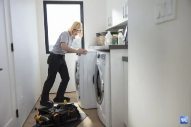 Technician in glasses wearing a light grey polo shirt and dark trousers leaning over a washing machine in a modern laundry room.