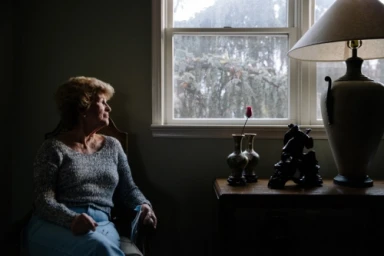 Elderly woman seated by a window inside a living room, photographed by Augustus Butera