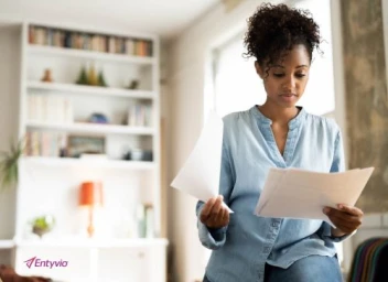 Black woman in a light blue denim shirt reviewing documents indoors with a bookshelf in the background.