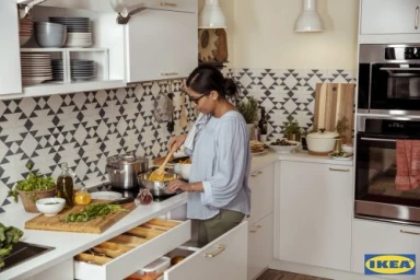 Woman in a light blue blouse cooking in a modern white kitchen with geometric tile backsplash, photographed by Augustus Butera