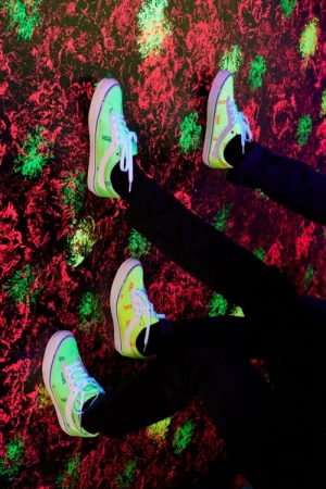 Two pairs of neon-green Vans sneakers are positioned on a red and black textured background in a vibrant, visually striking composition.