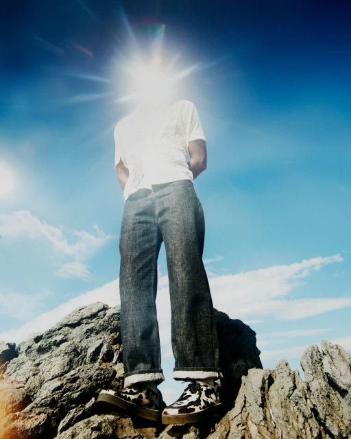 Person standing on rock formation with sun behind their head, wearing jeans, white shirt and animal print shoes against sky.