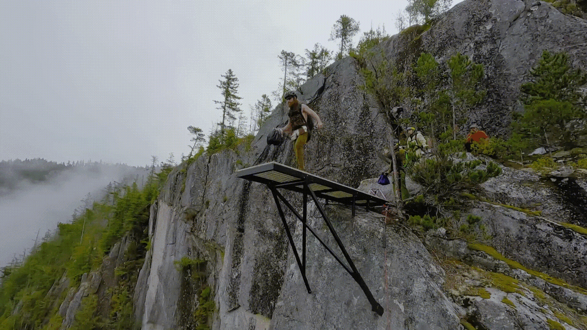An adult male in outdoor sportswear stands on a platform atop a cliff in a mountain environment with some other people in the background.