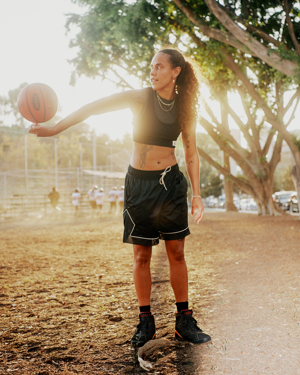 Athletic woman outdoors balancing a basketball on her finger while wearing a black sports bra and shorts with bright sunlight.