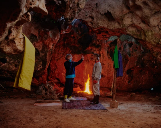 Two people in a cave stand near a fire in the center; one poses with a phone for a photo while colorful garments hang nearby.