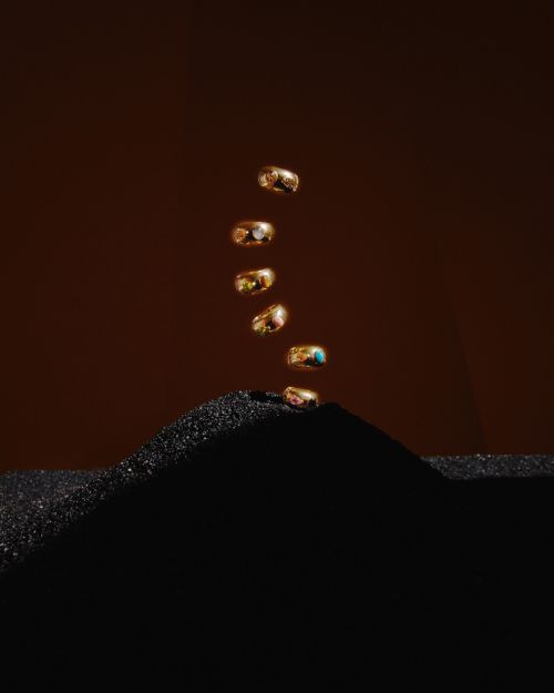 Seven gold rings with colorful gems appear to levitate on top of a black sand dune against a dark brown backdrop.