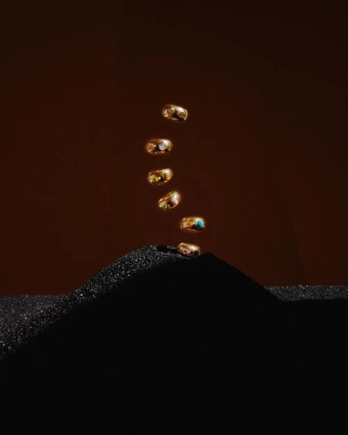Seven gold rings with colorful gems appear to levitate on top of a black sand dune against a dark brown backdrop.