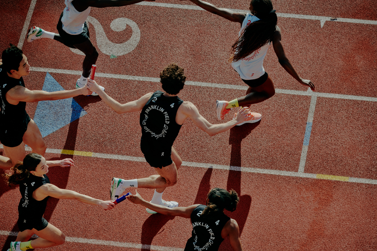 Top-down shot of a relay race with athletes passing batons on a red track, showcasing teamwork and athletic competition on a sunny day.