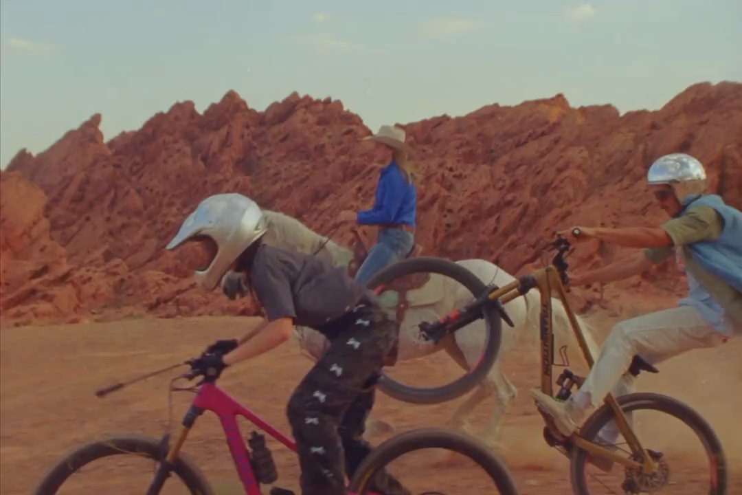 Three people cycling and riding a horse through a desert landscape with red rock formations under a clear sky, active pursuits.
