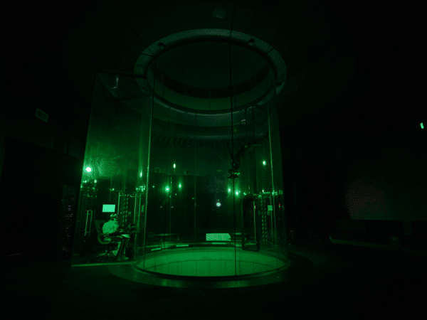 Indoor skydive tunnel with person visible inside, instructor seated outside tunnel. Green-toned lighting inside a dark environment.