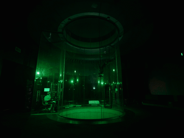 Indoor skydive tunnel with person visible inside, instructor seated outside tunnel. Green-toned lighting inside a dark environment.