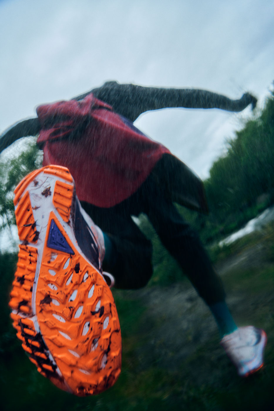 Close-up of a running shoe's orange sole in mid-stride with a person in motion on a trail. Action shot captures speed and outdoor activity.