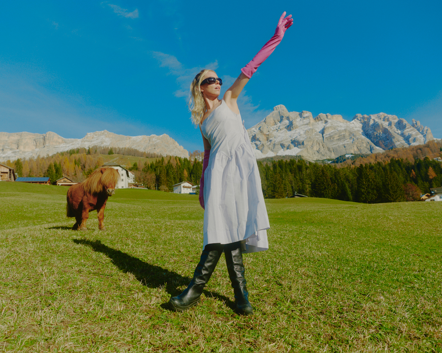 A blonde woman in a white dress poses in a green field with a miniature horse, mountains, trees, and a bright blue sky in the background.