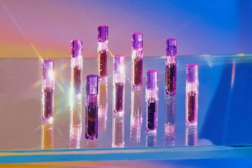 Nine purple-capped lip glosses stand on a reflective surface with a blue and orange gradient background, creating a sleek beauty product display.