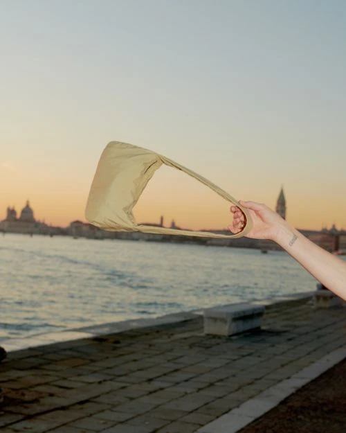 Beige shoulder bag held up by a person's hand outdoors against a sunset over water and a distant city skyline.